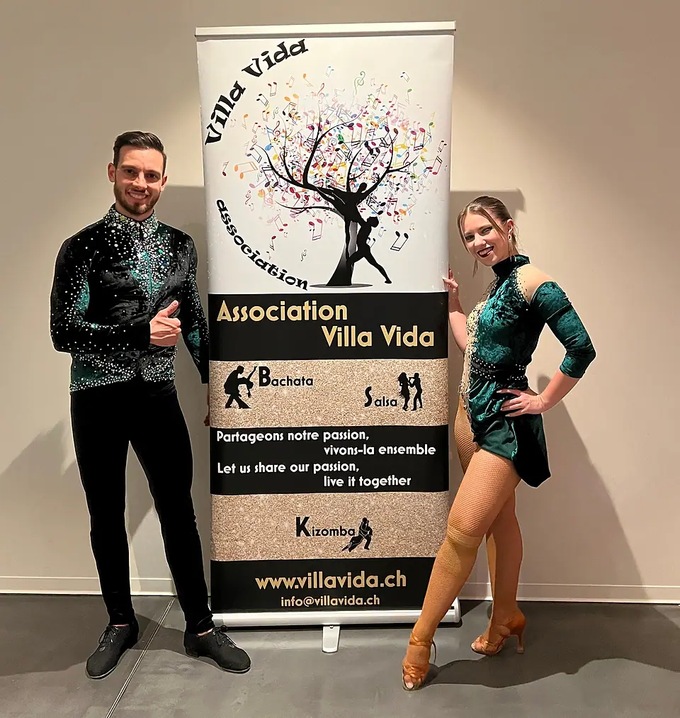 Un danseur et une danseuse en costumes de scène verts, de part et d’autre du roll-up Villa Vida.