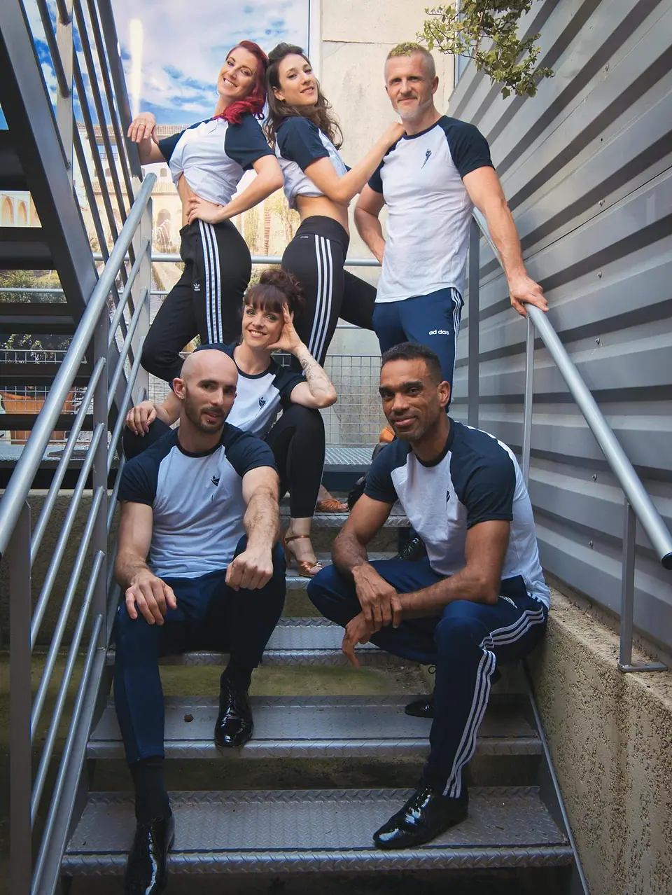 Six danseuses et danseurs en tenues assorties, assis et debout dans un escalier métallique.
